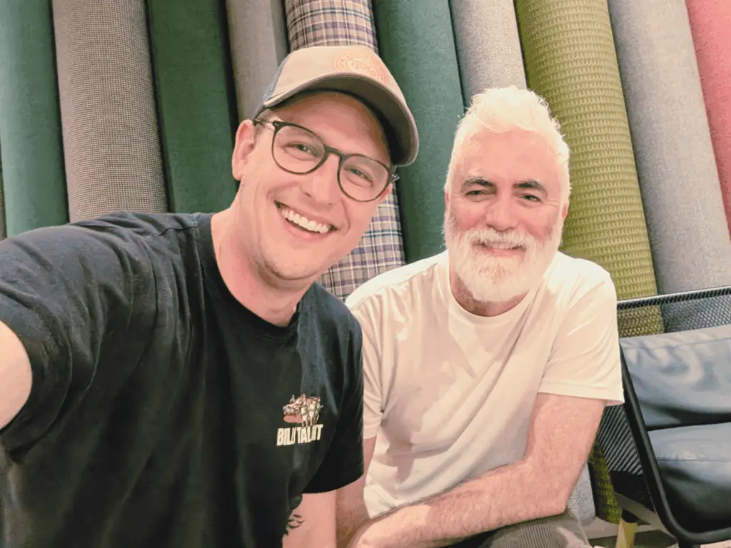Two men selfie and smiling one wearing hat the other has a white beard with a coloured background.