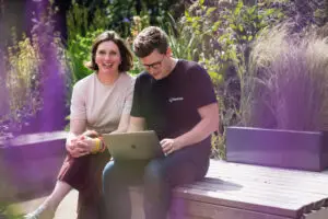 Woman smiles at camera while man works on computer in a garden