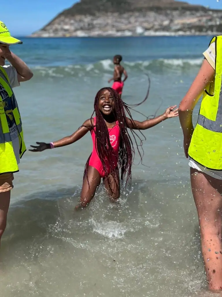 A smiling kid in the sea with long braided hair taking part in the Darcey Sunshine Swim programme with instructors around her