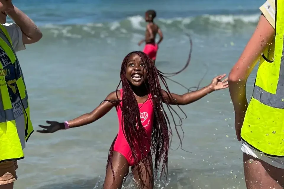 A smiling kid in the sea with long braided hair taking part in the Darcey Sunshine Swim programme with instructors around her