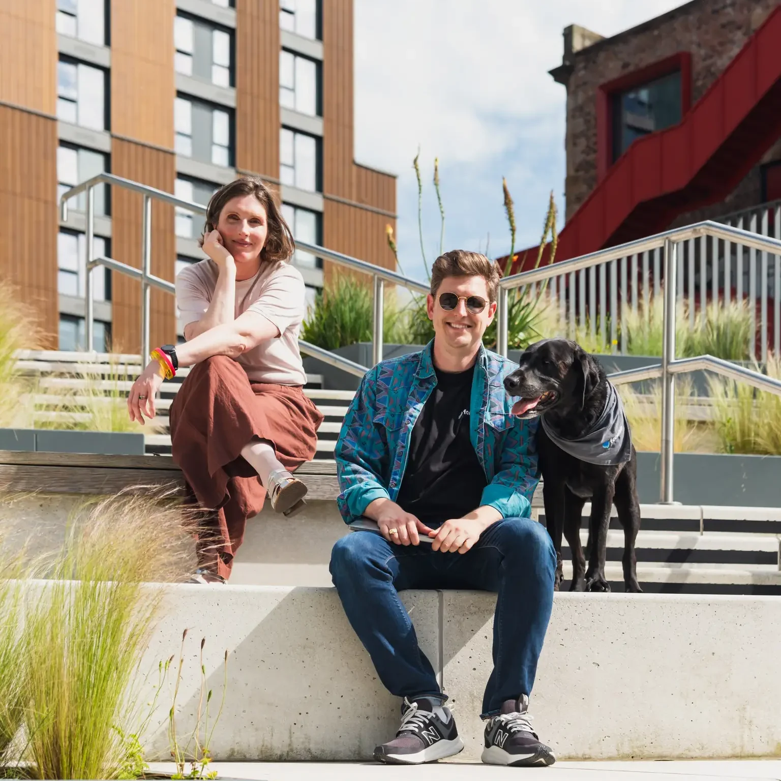 Lady and man with sunglasses it on steps looking at camera with black dog beside them